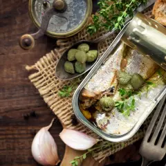 Une boîte ouverte de la sardine, garnie de câpres et de thym, sur un fond de bois rustique avec de l'ail et des toasts.