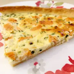 Une part de quiche au saumon fumé, garnie d'herbes fraîches, sur une assiette décorée de fleurs rouges et vertes.