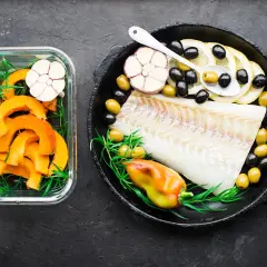 Filet de cabillaud poisson cru avec citrons, olives, ail et poivron dans une poêle, à côté de courge coupée en dés dans un récipient en verre.