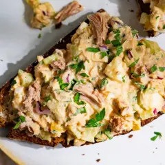 Une tranche de pain noir garnie d'une généreuse salade de thon crémeuse, avec des morceaux d'œuf dur, de céleri et d'oignon rouge, parsemée de persil frais.