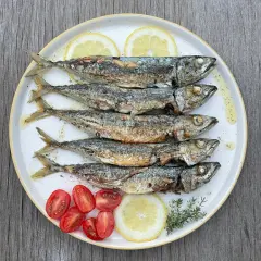 Cinq chinchards poissons grillés, servis sur une assiette avec des quartiers de citron, des tomates cerises et des herbes.
