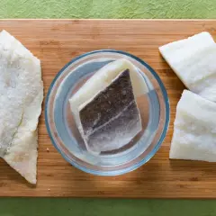 Morue salée prête pour une recette. Des morceaux de poisson sont disposés sur une planche en bois, certains dans un bol d'eau.