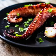 Tentacules de poulpe grillés, parsemés de persil frais, sur une assiette noire. La cuisson du poulpe révèle sa texture tendre.