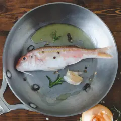 Un poisson rouge prêt à être cuisiné à la poêle avec de l'ail, du romarin et des grains de poivre sur une planche en bois.