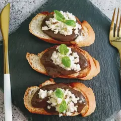 Trois toasts de pain grillé garnis d'une pâte foncée, de fromage blanc émietté et de feuilles de menthe. Une présentation élégante pour une tapenade de la mer revisitée.