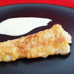 Un morceau de lingue poisson pané, doré et croustillant, servi avec une sauce blanche crémeuse sur une assiette noire.