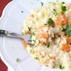Un crémeux risotto fruits de mer, garni de crevettes roses, persil frais et zeste de citron. Une fourchette est plantée dans ce plat savoureux.