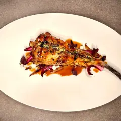 Un plat de lotte au barbecue, laqué et garni d'herbes, servi sur un lit de légumes rouges et blancs.