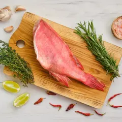 Un sébaste poisson cru, prêt à être cuisiné avec ail, thym, romarin, piments et sel rose sur une planche en bois.