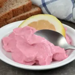 Une cuillère sert une portion de tarama rose crémeux, garnie d'une tranche de citron. Du pain grillé est visible en arrière-plan. Une recette simple pour un apéritif savoureux.