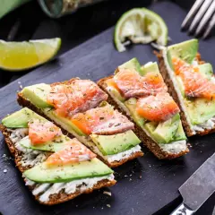 Quatre toasts avocat saumon, garniture crémeuse, herbes et quartiers de citron vert sur planche noire.