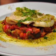 Un délicieux dos de cabillaud servi sur un lit de légumes méditerranéens, arrosé d'une sauce verte et d'huile d'olive.