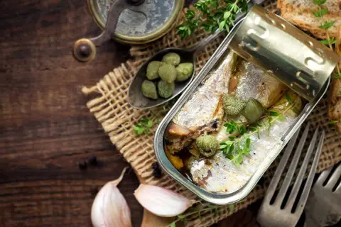 Une boîte ouverte de la sardine, garnie de câpres et de thym, sur un fond de bois rustique avec de l'ail et des toasts.