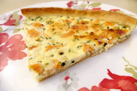 Une part de quiche au saumon fumé, garnie d'herbes fraîches, sur une assiette décorée de fleurs rouges et vertes.
