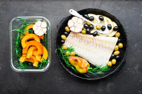 Filet de cabillaud poisson cru avec citrons, olives, ail et poivron dans une poêle, à côté de courge coupée en dés dans un récipient en verre.