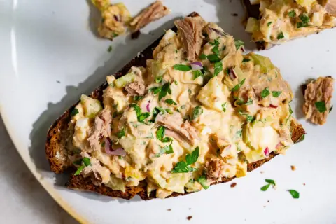 Une tranche de pain noir garnie d'une généreuse salade de thon crémeuse, avec des morceaux d'œuf dur, de céleri et d'oignon rouge, parsemée de persil frais.