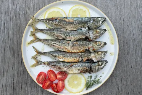Cinq chinchards poissons grillés, servis sur une assiette avec des quartiers de citron, des tomates cerises et des herbes.
