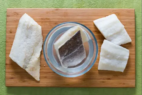 Morue salée prête pour une recette. Des morceaux de poisson sont disposés sur une planche en bois, certains dans un bol d'eau.