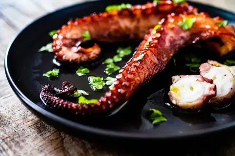 Tentacules de poulpe grillés, parsemés de persil frais, sur une assiette noire. La cuisson du poulpe révèle sa texture tendre.
