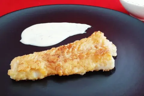 Un morceau de lingue poisson pané, doré et croustillant, servi avec une sauce blanche crémeuse sur une assiette noire.