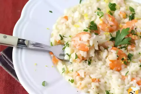 Un crémeux risotto fruits de mer, garni de crevettes roses, persil frais et zeste de citron. Une fourchette est plantée dans ce plat savoureux.