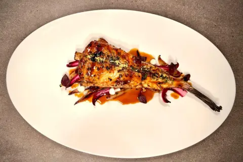 Un plat de lotte au barbecue, laqué et garni d'herbes, servi sur un lit de légumes rouges et blancs.