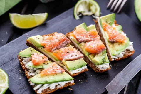 Quatre toasts avocat saumon, garniture crémeuse, herbes et quartiers de citron vert sur planche noire.