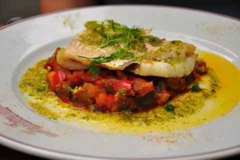 Un délicieux dos de cabillaud servi sur un lit de légumes méditerranéens, arrosé d'une sauce verte et d'huile d'olive.