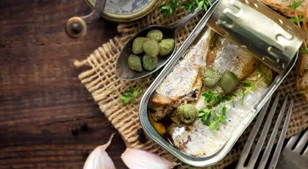 Une boîte ouverte de la sardine, garnie de câpres et de thym, sur un fond de bois rustique avec de l'ail et des toasts.