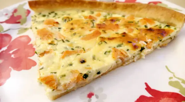 Une part de quiche au saumon fumé, garnie d'herbes fraîches, sur une assiette décorée de fleurs rouges et vertes.