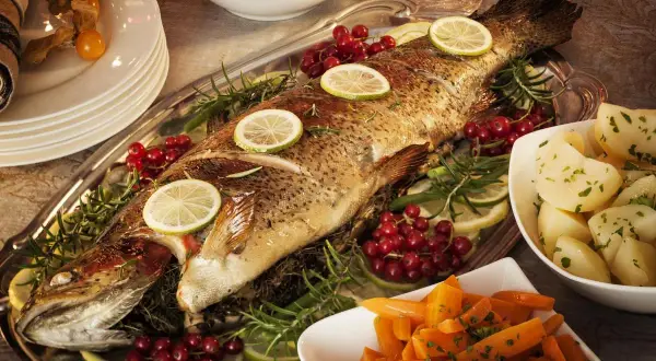 Un poisson entier, un turbot, est servi sur un plateau avec des rondelles de citron, des airelles et du romarin. Des pommes de terre et des carottes sont disposées sur les côtés.
