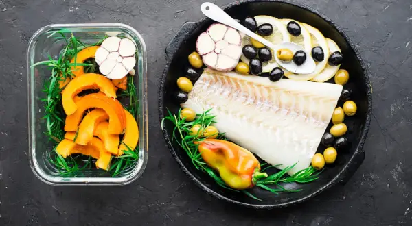 Filet de cabillaud poisson cru avec citrons, olives, ail et poivron dans une poêle, à côté de courge coupée en dés dans un récipient en verre.