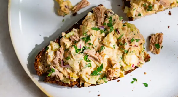 Une tranche de pain noir garnie d'une généreuse salade de thon crémeuse, avec des morceaux d'œuf dur, de céleri et d'oignon rouge, parsemée de persil frais.