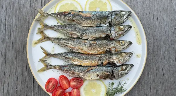 Cinq chinchards poissons grillés, servis sur une assiette avec des quartiers de citron, des tomates cerises et des herbes.