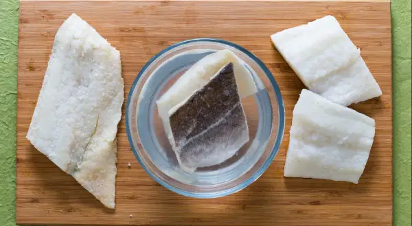 Morue salée prête pour une recette. Des morceaux de poisson sont disposés sur une planche en bois, certains dans un bol d'eau.