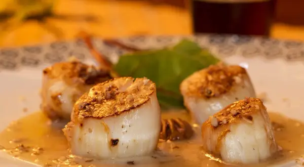 Quatre noix de Saint-Jacques poêlées, dorées et nappées d'une sauce onctueuse, sur une assiette décorée.