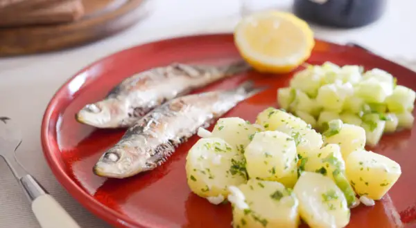 Deux sardines grillées sur une assiette rouge, accompagnées de pommes de terre persillade et d'une salade de concombre. Un vrai délice de sardine barbecue !