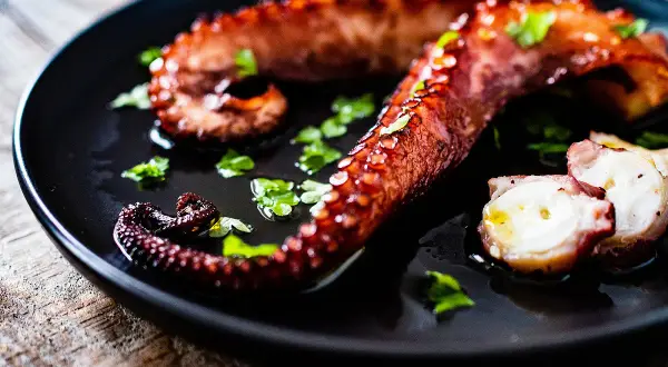 Tentacules de poulpe grillés, parsemés de persil frais, sur une assiette noire. La cuisson du poulpe révèle sa texture tendre.