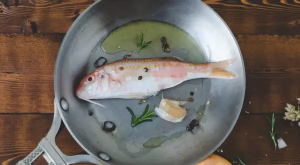 Un poisson rouge prêt à être cuisiné à la poêle avec de l'ail, du romarin et des grains de poivre sur une planche en bois.
