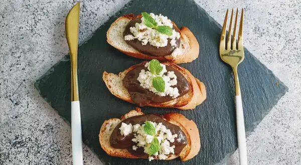 Trois toasts de pain grillé garnis d'une pâte foncée, de fromage blanc émietté et de feuilles de menthe. Une présentation élégante pour une tapenade de la mer revisitée.