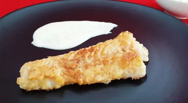 Un morceau de lingue poisson pané, doré et croustillant, servi avec une sauce blanche crémeuse sur une assiette noire.