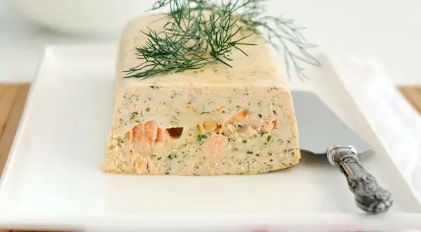 Terrine de saumon frais et fumé garnie d'aneth sur une assiette blanche avec un couteau.