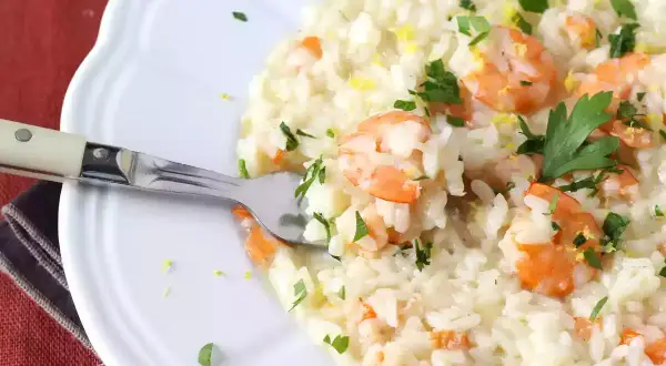 Un crémeux risotto fruits de mer, garni de crevettes roses, persil frais et zeste de citron. Une fourchette est plantée dans ce plat savoureux.