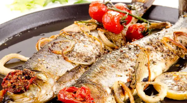 Deux poissons entiers, le hareng, rôtis avec des tomates cerises, des oignons et des rondelles de citron sur un plat noir.