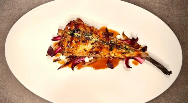 Un plat de lotte au barbecue, laqué et garni d'herbes, servi sur un lit de légumes rouges et blancs.