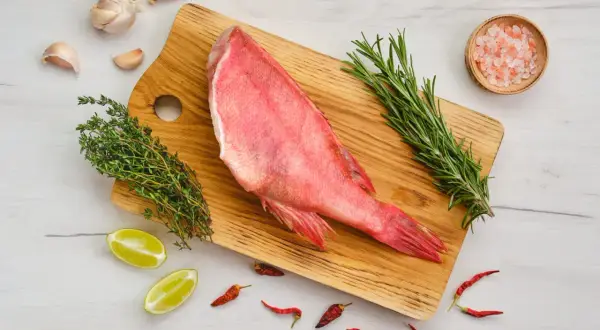Un sébaste poisson cru, prêt à être cuisiné avec ail, thym, romarin, piments et sel rose sur une planche en bois.