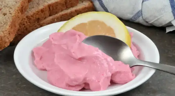 Une cuillère sert une portion de tarama rose crémeux, garnie d'une tranche de citron. Du pain grillé est visible en arrière-plan. Une recette simple pour un apéritif savoureux.