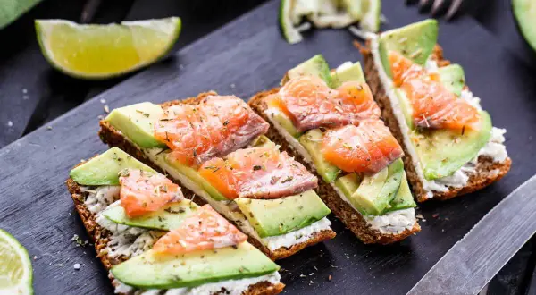 Quatre toasts avocat saumon, garniture crémeuse, herbes et quartiers de citron vert sur planche noire.