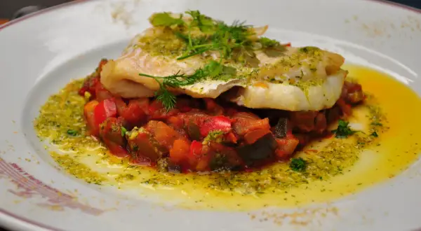 Un délicieux dos de cabillaud servi sur un lit de légumes méditerranéens, arrosé d'une sauce verte et d'huile d'olive.