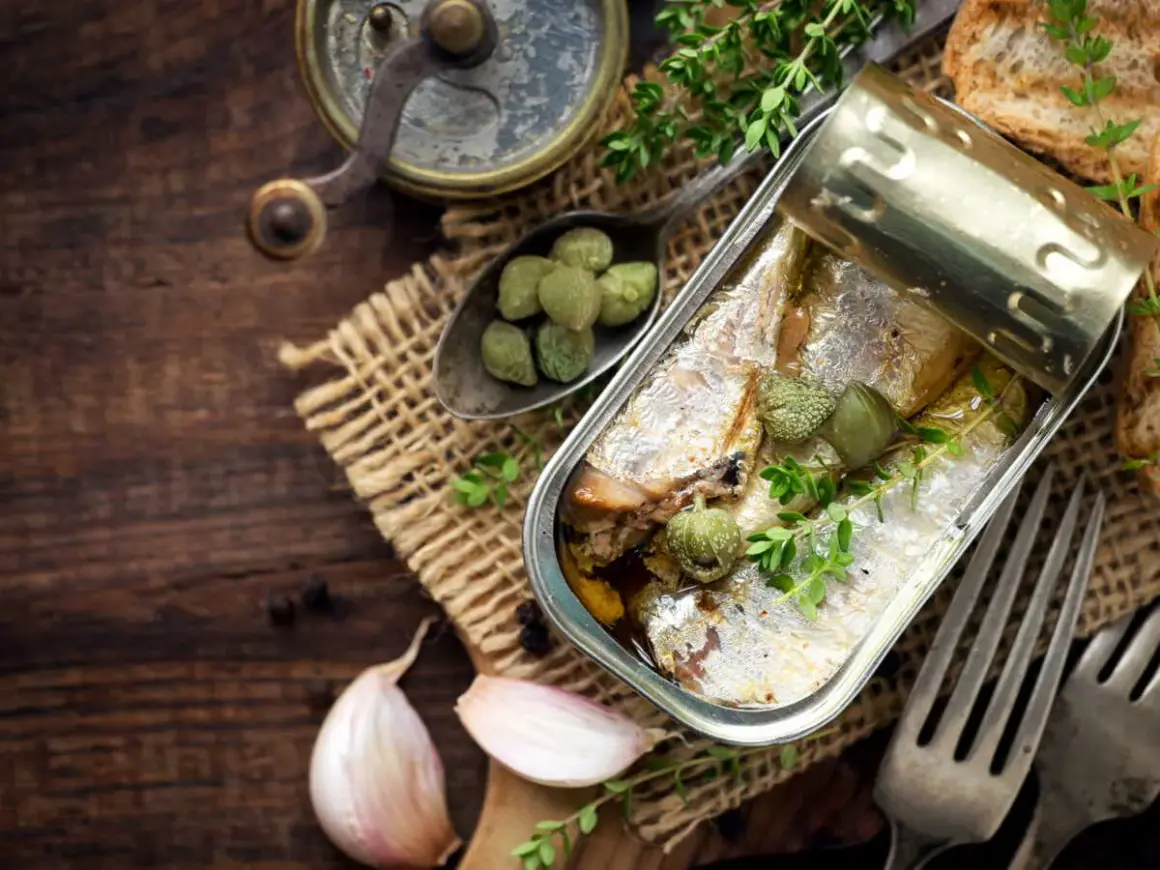 Une boîte ouverte de la sardine, garnie de câpres et de thym, sur un fond de bois rustique avec de l'ail et des toasts.